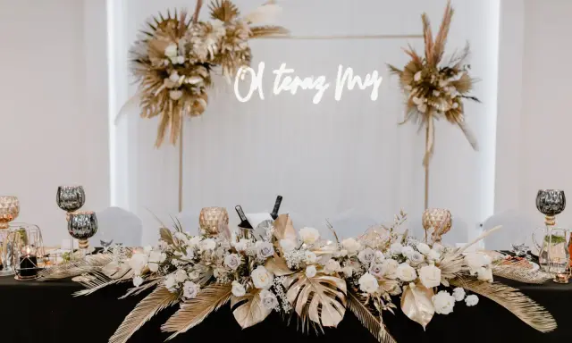 Elegancka ścianka za młodymi boho z napisem "O, teraz My". Dekoracja stołu z bukietami kwiatów, złotymi liśćmi i świecami.