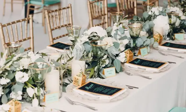 Elegancko nakryty stół z menu i kwiatami. Złote krzesła i zielone kieliszki tworzą atmosferę uroczystego obiadu weselnego zamiast wesela.
