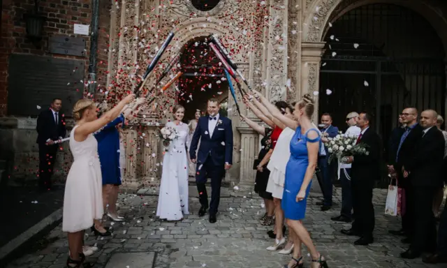 Szczęśliwa para młoda wychodzi z kościoła, obsypana płatkami. Goście z radością świętują wyjście z kościoła ślub.