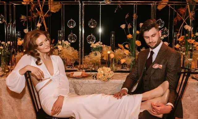 Para młoda przy stole, ona w białej sukni, on w brązowej marynarce. Romantyczna atmosfera, kwiaty i świece.