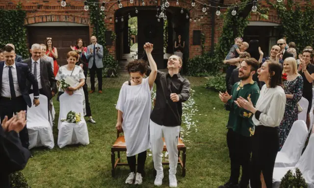 Para młoda świętuje ślub cywilny na luzie, trzymając się za ręce. Goście klaszczą, ciesząc się radosnym momentem.