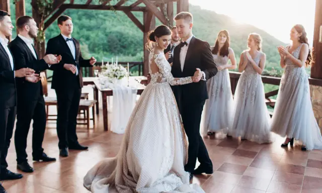 Para Młoda tańczy swój pierwszy taniec. Panna Młoda w pięknej sukni, Pan Młody w eleganckim garniturze. Druhny i świadkowie patrzą z uśmiechem.