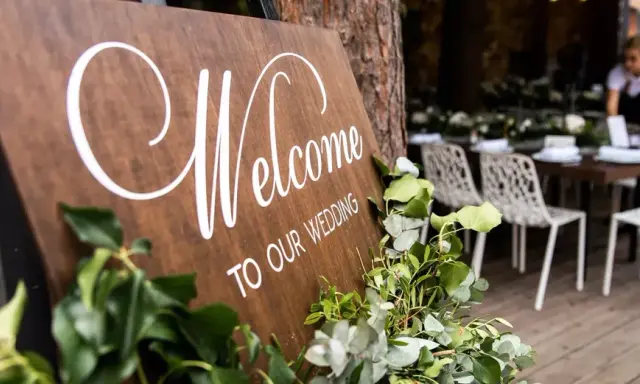 Drewniana tablica "Welcome to Our Wedding" ozdobiona zielenią. W tle widać przygotowane stoły i krzesła, a także obsługę. Opłata ZAIKS za muzykę na weselu.