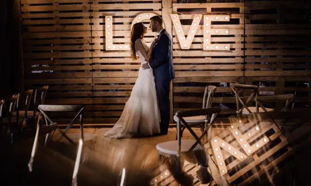 Para młoda w objęciach na tle napisu "LOVE" z żarówek. Piękny plener ślubny z drewnianą ścianą i krzesłami.