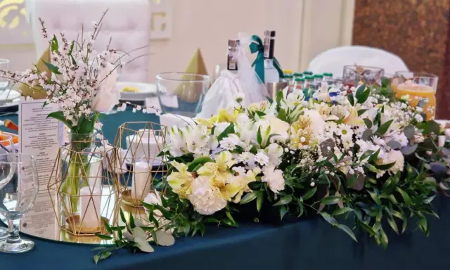 Elegancki pomysł na wesele: stół udekorowany bukietami białych i żółtych kwiatów, świecami w złotych lampionach i butelkami z napojami.