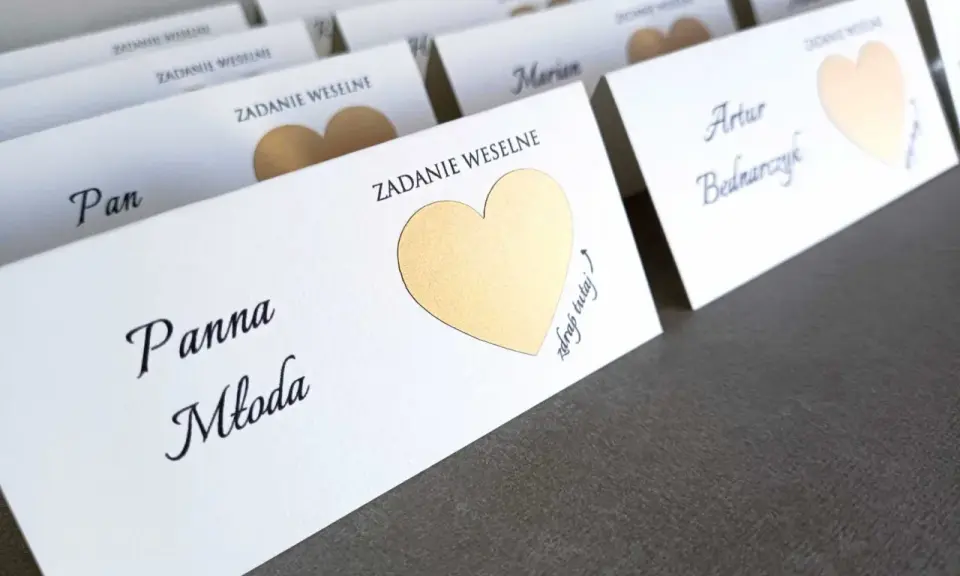 Białe karty z imionami gości i złotymi serduszkami kryją śmieszne zadania dla gości weselnych.