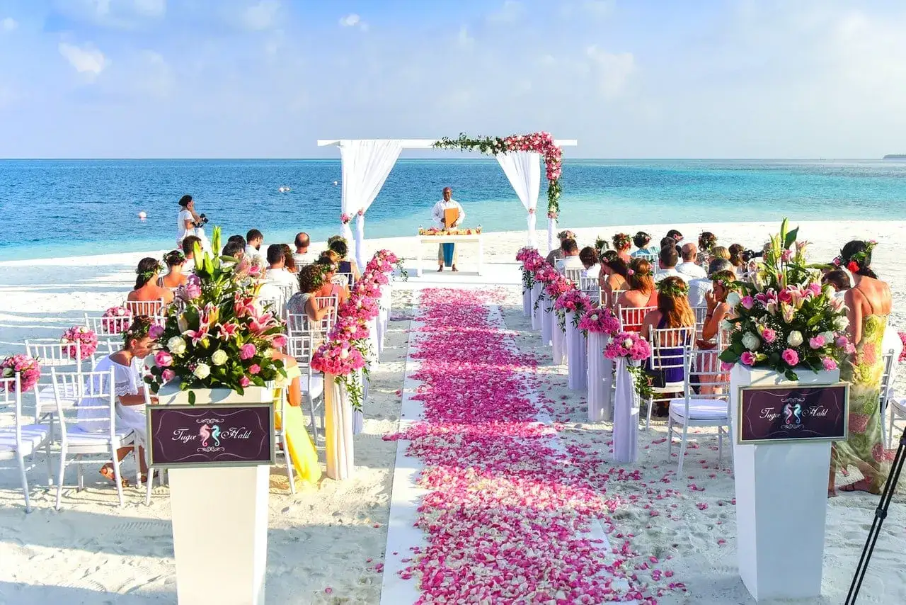 Nietypowe miejsca na wesele: romantyczna ceremonia na plaży z widokiem na ocean, udekorowana białymi krzesłami, różowymi kwiatami i dywanem z płatków.