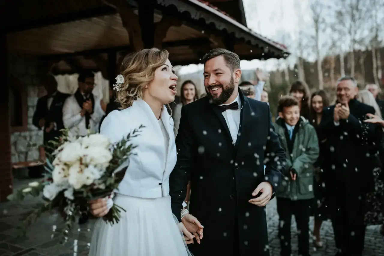 Szczęśliwa para młoda, trzymająca bukiet, wychodzi na śnieżny ślub bez zapowiedzi. Goście sypią płatkami.