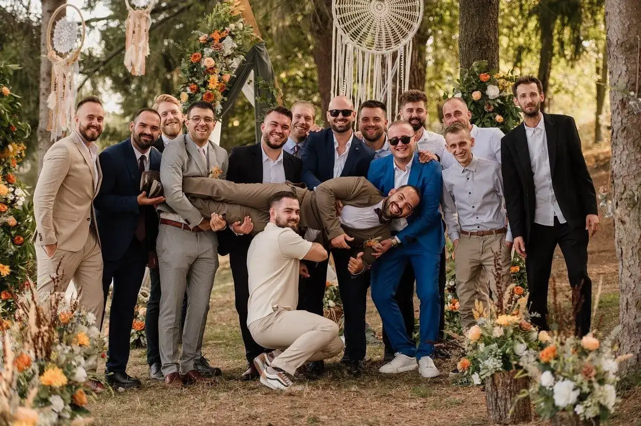 Panowie świętują ślub symboliczny. Jeden z nich jest niesiony na rękach przez przyjaciół, wszyscy uśmiechnięci i pełni radości.
