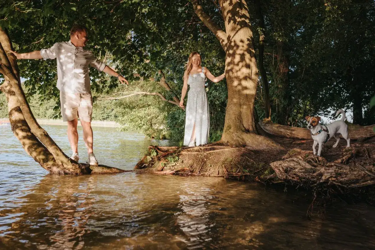 Para sesji ślubnej: on na drzewie nad wodą, ona na brzegu, piesek czeka. Idealne pytania do fotografa ślubnego o takie ujęcia.