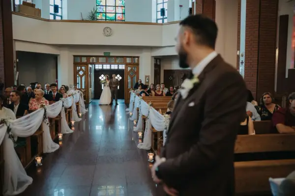 Panna młoda z ojcem idzie do ołtarza. W kościele panują uroczyste warunki ślubu kościelnego, z gośćmi siedzącymi w ławkach.